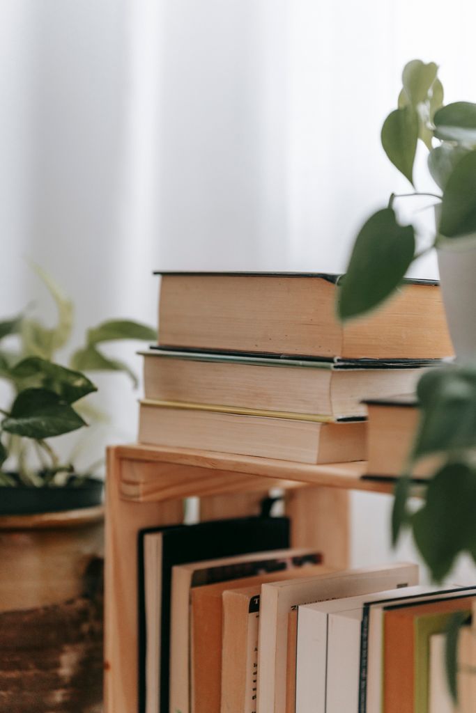 Stack of various books arranged on wooden shelf near green potted plants in light apartment